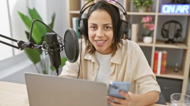 İspanyol kadın podcaster mikrofon ve akıllı telefonla bir radyo stüdyosunda kameraya gülümsüyor..