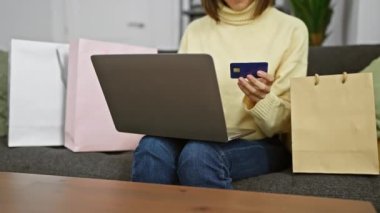Genç bir kadın alışveriş torbalarının arasında kredi kartı ve laptopuyla internetten alışveriş yapıyor..