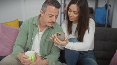 Akıllı telefonlu sevgi dolu bir çift modern bir oturma odasında rahatça ev hayatını canlandırıyor..