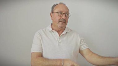 Gözlüklü mutlu, orta yaşlı bir adam kameraya gülümseyen beyaz bir arkaplanı işaret ediyor.