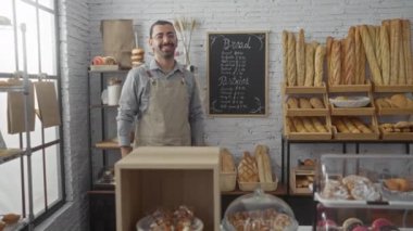 Ekmek ve pastalarla çevrili fırında duran İspanyol bıyıklı adam. Parlak, kapalı bir yerde gülümsüyor, içi fırınlanmış mallar ve karatahta menüleriyle dolu bir mağaza.