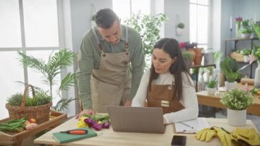 Canlı lalelerin arasında, bir adam ve bir kadın, önlüklü, kapalı bir çiçekçide dizüstü bilgisayara danışıyorlar..