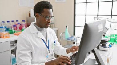 Profesyonel bir Afrikalı, bilimsel ekipmanlarla çevrili bir laboratuvar ortamında bilgisayar başında dikkatle çalışır..