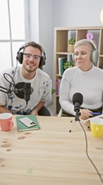 Parlak bir podcast stüdyosunda kulaklık takan bir kadın ve bir erkek mikrofonlara konuşuyor..