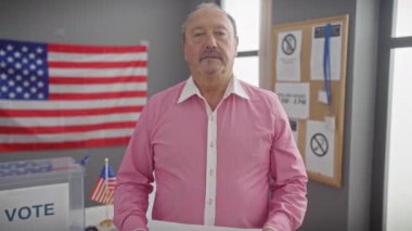 Pembe gömlekli olgun bir adam, arka planda Amerikan bayrağı olan bir seçim merkezinde 