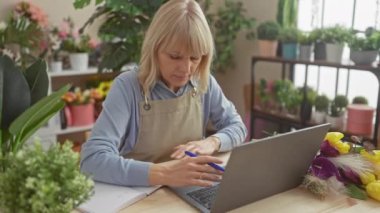 Önlüklü sarışın bir kadın dizüstü bilgisayarın yanındaki defterde yazıyor. Etrafı çiçekçiyle dolu..