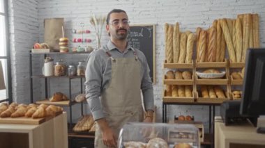 Kapalı bir fırında kolları çapraz duran genç erkek fırıncı çeşitli ekmek türleri ve raflarda sergilenen hamur işleri ile çevrili.