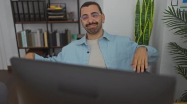 Ofis ortamında bıyıklı genç bir adam, bilgisayarla çalışıyor, kapalı mekan bitkileri ve ofis malzemeleriyle çevrili olumlu duyguları ifade ediyor ve esnetiyor..