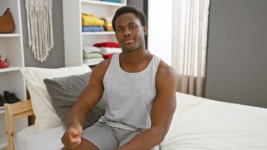 Neşeli, Afro-Amerikalı genç adam rahat yatak odasında baş parmak hareketi yapıyor, rahat pijamalarıyla başarıyı onaylıyor, güzel bir gülümsemeyle kendinden emin bir poz veriyor..