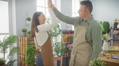 Çiçekçi dükkanında gülümseyen bir kadın ve erkek beşlik çakıyor, ikisi de önlük giyiyor, takım çalışmasını ve ortaklığı simgeliyor..