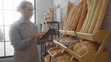 Fırında elinde tablet olan genç bir adam çeşitli taze ekmek somunlarıyla çevrili ürünleri inceliyor.