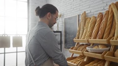 İspanyol bir adam pastanede çalışıyor ahşap raflarda ekmek düzenlerken kapalı bir ortamda gülümsüyor.