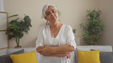 Oturma odasında oturan gri saçlı, beyaz bluzlu ve kollarını çapraz bağlayan olgun bir kadın rahat bir evde bitkilerle ve sarı yastıklarla çevrili.