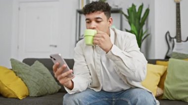 Genç adam kahve içiyor ve modern bir oturma odasında akıllı telefon kullanıyor..