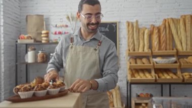 Yakışıklı bir adam pastanede çalışıyor, gülümseyerek hamur işleri hazırlıyor. Etrafı ev ortamında taze ekmekle çevrili.