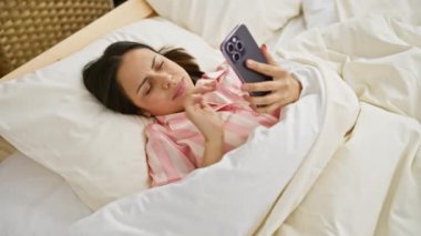 Pembe pijamalı genç bir İspanyol kadın, rahat bir yatak odası ortamında akıllı telefonuyla iletişim kuruyor.
