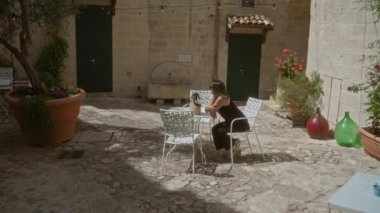 Genç İspanyol kadın, tarihi eski bir kasaba olan Mata, Basilicata, İtalya 'da güneşli bir günün tadını çıkarıyor. Açık bir kafede oturmuş, saksı bitkileri, canlı çiçekler ve antika mimari ile süslenmiş bir şekilde.. 