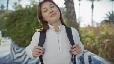 Sırt çantalı neşeli genç bir kadın güneşli bir şehir parkının önünde duruyor..