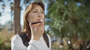 Yeşilliklerle çevrili genç, beyaz bir kadın telefonuna sesli mesaj kaydediyor..