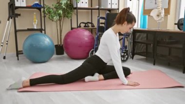 Genç bir İspanyol kadın, rehabilitasyon salonunda egzersiz topları ve tıbbi malzemelerle yoga minderinin üzerine uzanıyor..