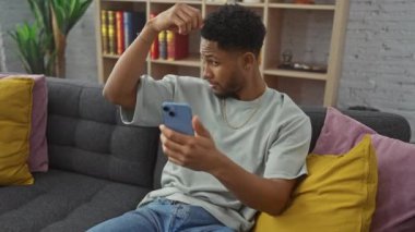 Kafası karışmış genç bir zenci telefonunu renkli yastıklı gri bir koltukta modern bir oturma odasında kontrol ediyor..