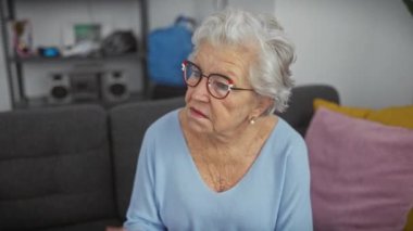 Gözlüklü yaşlı kadın, oturma odasında düşünceli bir şekilde oturuyor, yaşlı yaşam tarzını ve evde boş vakitlerini resmediyor..