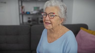 Modern bir oturma odasında gözlüklü neşeli yaşlı bir kadın samimi bir şekilde gülümsüyor, sıcaklığını ve rahatlığını sergiliyor..