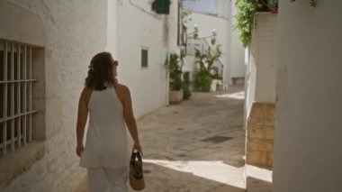 Genç bir İspanyol kadın Locorotondo, Puglia, İtalya sokaklarında yürüyor. Etrafı beyaz binalar ve canlı yeşilliklerle çevrili..