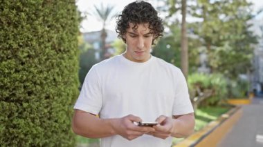Kıvırcık saçlı yakışıklı bir İspanyol, güneşli bir caddede dikilirken akıllı telefonunu kullanıyor.
