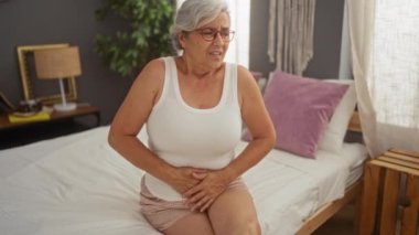 Yaşlı bir kadın, gri saçlı, yatak odasında bir yatakta oturuyor, karnını acıdan tutuyor..