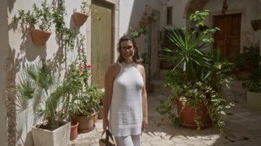 Beyaz elbiseli güzel bir İspanyol kadın güneşli bir günün keyfini çıkarırken İtalya 'da yemyeşil bitkiler ve eski kasaba mimarisi ile çevrili büyüleyici locorotondo, puglia sokaklarında dikiliyor..