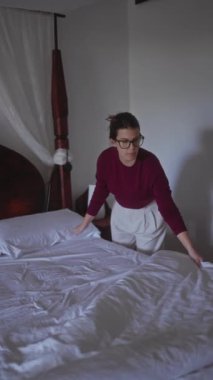 Odaklanmış bir kadın, yumuşak ışıklı bir yatak odasında beyaz bir yatak düzenler, ev ve sükunet hissi uyandırır..