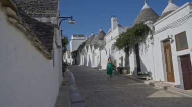 Yeşil elbiseli genç bir İspanyol kadın açık mavi gökyüzünün altında ikonik Trulli evlerle çevrili, İtalya 'nın büyüleyici eski Alberobello kasabasında bir kaldırım taşı caddesinde yürüyor..