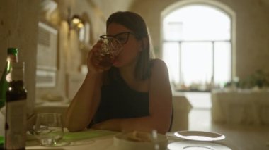 Genç İspanyol kadın, güzel aydınlatılmış bir İtalyan restoranında içkisini yudumlarken masada içkiler ve tabaklar ile rahatlatıcı bir ortam sergiliyor..