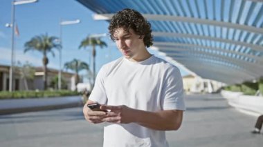 Genç İspanyol adam güneşli şehir ortamında palmiye ağaçlarıyla akıllı telefon kullanıyor.