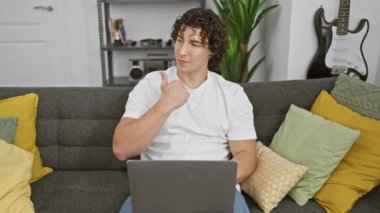 Neşeli, genç İspanyol bir adam, evinde koltukta rahatça oturuyor, dizüstü bilgisayardaki fikri işaret ediyor, ağzı açık gülümsüyor..