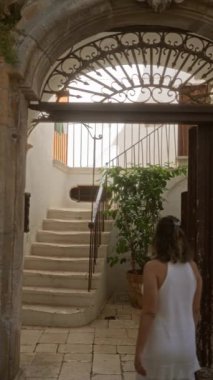 Beyaz elbiseli genç bir İspanyol kadın Locorotondo, Puglia, İtalya 'da süslü bir kemerin altındaki taş basamaklara tırmanıyor tarihi şehir sokaklarında ve Akdeniz mimarisinde..