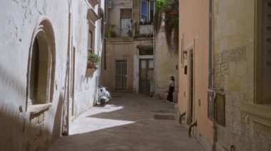 Bir kadın, Lecce, Puglia 'da dar, güneşli bir sokakta yürüyor. Eski binalar ve sokakta park etmiş bir scooter var..