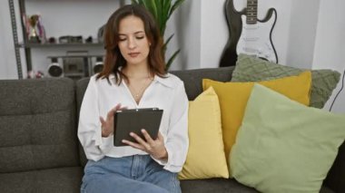 Odaklanmış İspanyol bir kadın rahatça döşenmiş oturma odasında tablet kullanıyor ve arka planda gitar var.