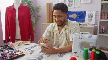 Etrafı kumaş, eskizler, dikiş makinesi ve mankenlerle çevrili renkli atölyelerde akıllı telefon kullanan gülen bir terzi.