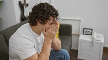 Endişeli, genç, İspanyol bir adam ellerini yüzünü kapatarak evde oturuyor, rahat bir oturma odasında stres yapıyor..