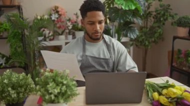 Afro-Amerikalı bir adam ev içi çiçekçide laptop ve renkli buketlerle doküman okuyor..