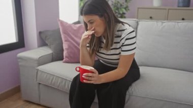 Esmer saçlı, Latin kökenli genç bir kadın. Oturma odasındaki kanepede oturuyor. Elinde kırmızı bir kupayla dalgın dalgın dalgın bakıyor..