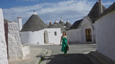 Yeşil elbiseli genç bir İspanyol kadın güneşli bir günde tarihi Alberobello, Puglia, İtalya 'da ikonik trulli evlerle dolu caddelerde yürüyor..