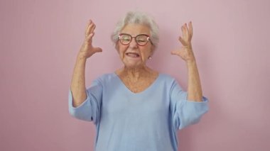 Gözlüklü öfkeli yaşlı kadın, gri saçlı, öfkeyle ayakta duran, bağıran, hayal kırıklığına uğrayan. Öfkeyle yükselen kollar pembe izole arka plan üzerine.