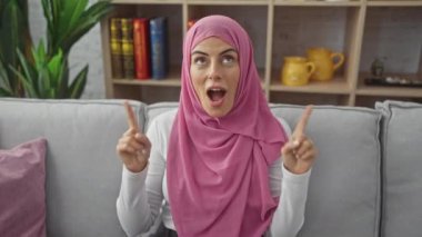 Büyülenmiş, tesettürlü genç İspanyol bir kadın, sevinçle evinin kanepesinde oturuyor, şaşırmış, ağzı açık, gözleri açık, parmakları şokunun noktasını işaret ediyor..