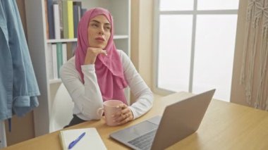 Ofiste dizüstü bilgisayarı, kitaplığı, fincanı ve pencere ışığıyla tesettürlü düşünceli bir kadın..