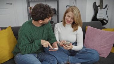 Modern bir oturma odasında rahat bir şekilde oturan bir erkek ve kadın, akıllı telefonlar üzerinden paylaşıyor ve iletişim kuruyor, gündelik ve bağlı bir yaşam tarzını somutlaştırıyor..