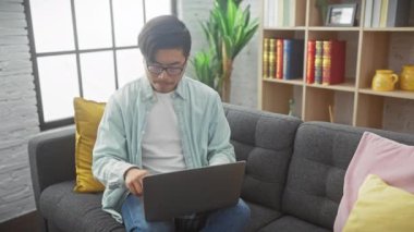 Genç Asyalı bir adam gri bir kanepede laptopuyla modern bir oturma odasında meditasyon yapıyor..