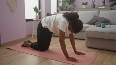 Genç bir Afrikalı Amerikalı kadın, rahat bir kanepe ve bitkilerle çevrili, rahat oturma odasında pembe bir paspasın üzerinde yoga egzersizi yapıyor..